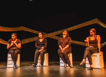 four women sit on blocks on stage with measuring tape in background